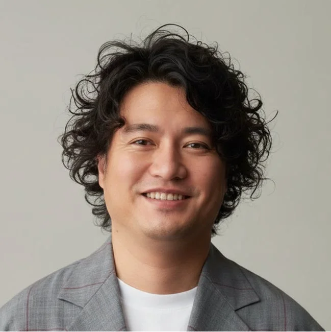 くるくるとした髪の男性がカメラに向かって笑顔を見せているポートレート写真です。グレーのジャケットを着用しており、背景はシンプルな単色です。