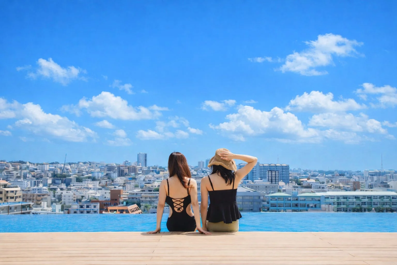 青い空と白い雲が広がる晴れた日、二人の女性がインフィニティプールの縁に座り、遠くの街並みを眺めている後ろ姿の画像です。リゾート感あふれる開放的な雰囲気が伝わります。
