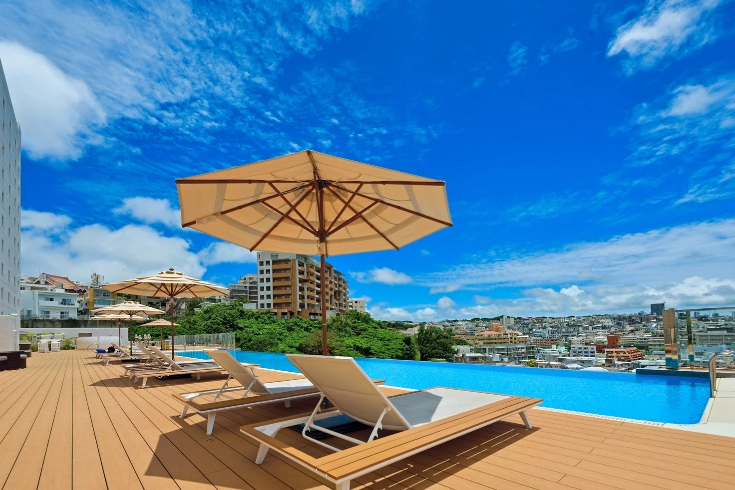 青い空と白い雲が広がる開放的な屋外プールサイドの風景です。木製のデッキには快適そうなラウンジチェアとパラソルが並び、遠くには市街地の景色が広がっています。リラックスできるリゾートのような雰囲気です。