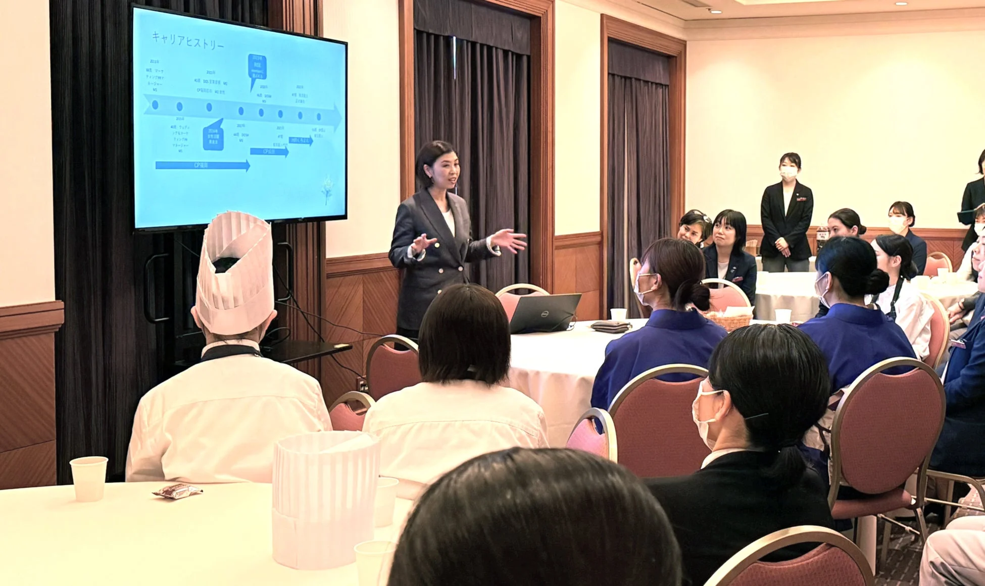会議室で女性が「キャリアヒストリー」と題されたスライドを使ってプレゼンテーションを行っています。聴講者には制服やコック帽を着用した多様な人々がおり、熱心に耳を傾けている研修やセミナーの様子です。