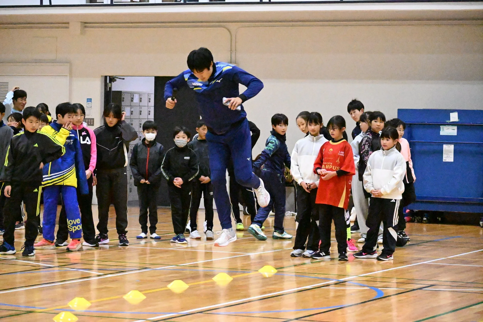 体育館で、ミズノのウェアを着た男性が子供たちの前でジャンプしている。子供たちは熱心にその様子を見守っており、スポーツイベントや教室の一幕のようだ。