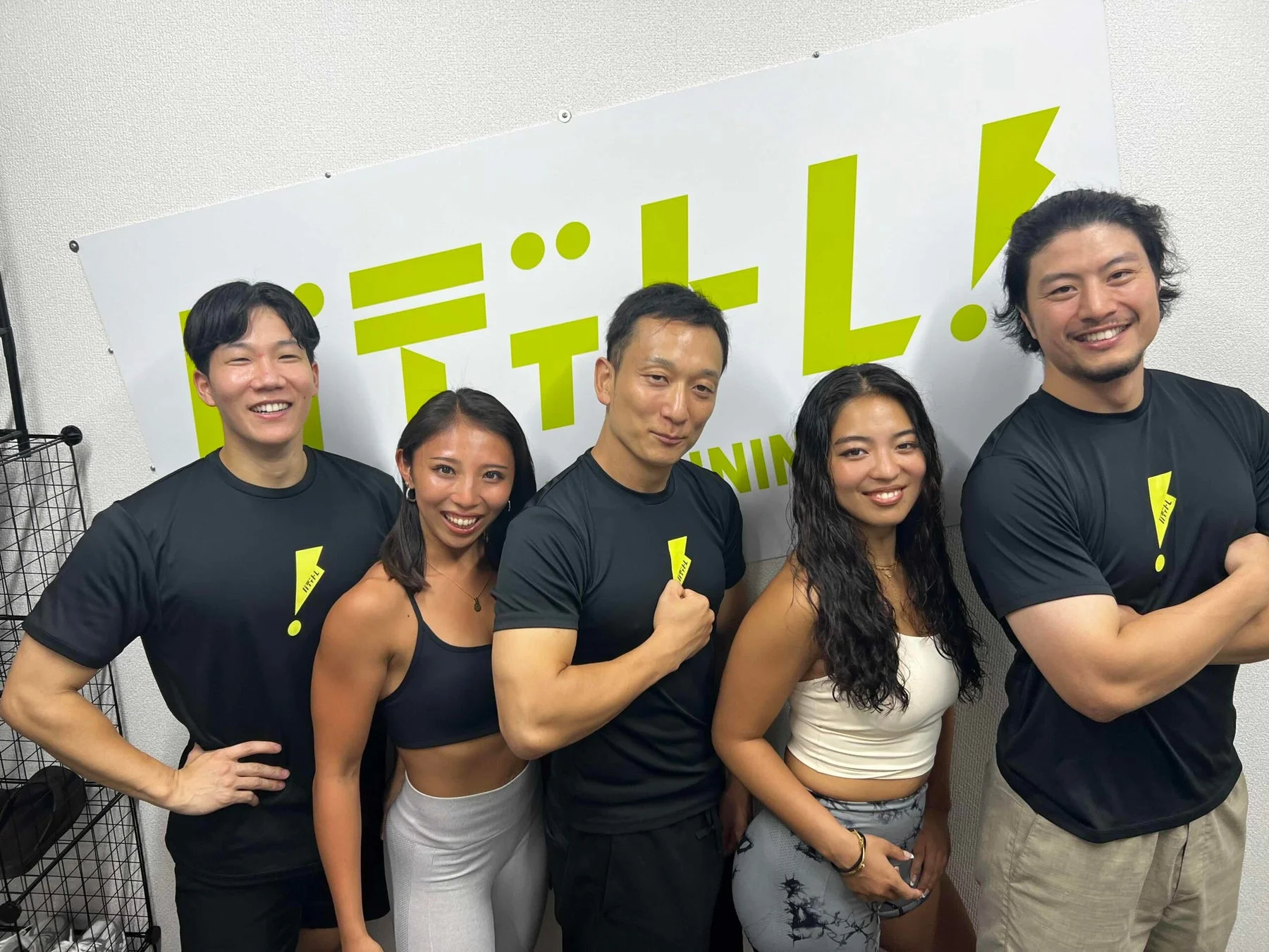 フィットネスジムで笑顔の男女5人がスポーツウェア姿で並んでポーズをとっています。