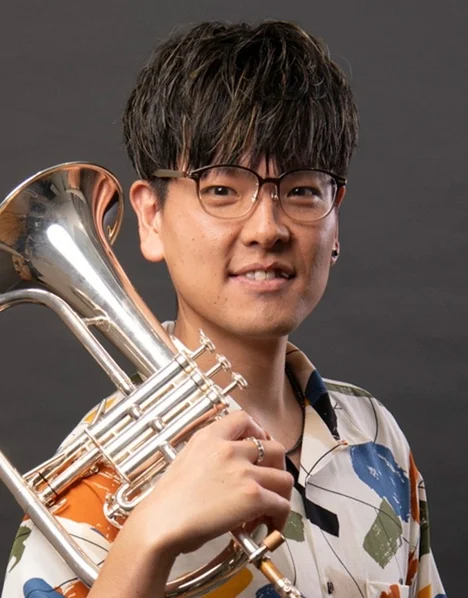 眼鏡をかけた男性がユーフォニアムのような金管楽器を抱え、笑顔で写っています。