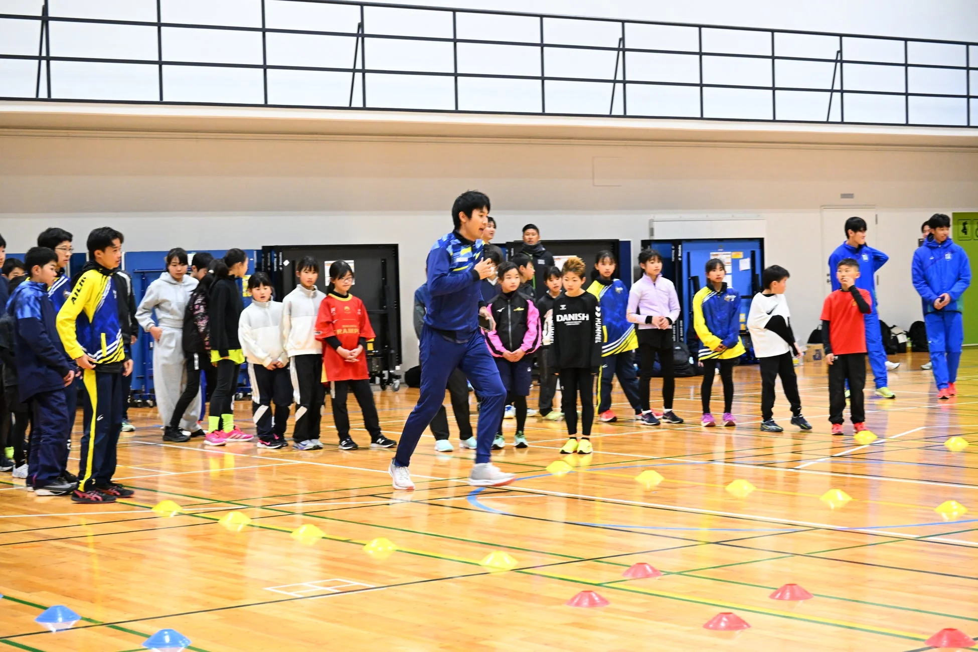 体育館で、多くの子どもたちが指導者の指示を聞きながらスポーツトレーニングをしている様子です。床にはカラフルなコーンが並べられています。