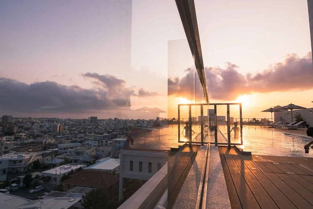 夕焼けに染まる空の下、都市のパノラマが広がる屋上プールで人々がくつろいでいる様子。美しい夕暮れの景色とリゾート感が漂う一枚です。