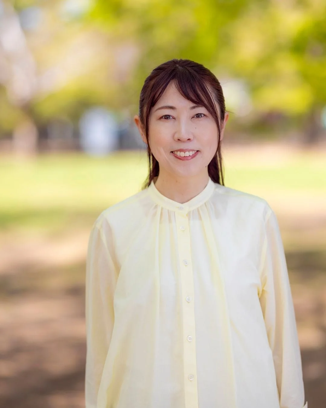 緑豊かな公園のような屋外で笑顔の女性がこちらを見て立っているポートレート写真