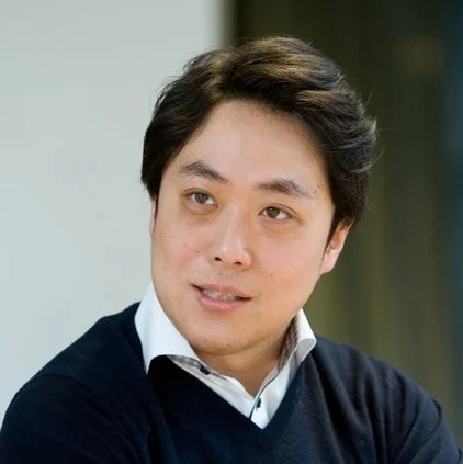 黒いVネックセーターと白いシャツを着用したアジア人男性のポートレートです。彼はカメラから少し目線を外し、穏やかな笑顔を浮かべています。背景はぼかされており、屋内であることが伺えます。