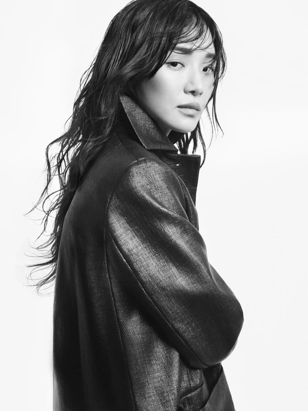 モノクロで撮影されたアジア人女性のポートレート。濡れたような長い髪と黒いジャケットが特徴的で、真剣な表情でカメラを見つめている。白背景が被写体を引き立てている。