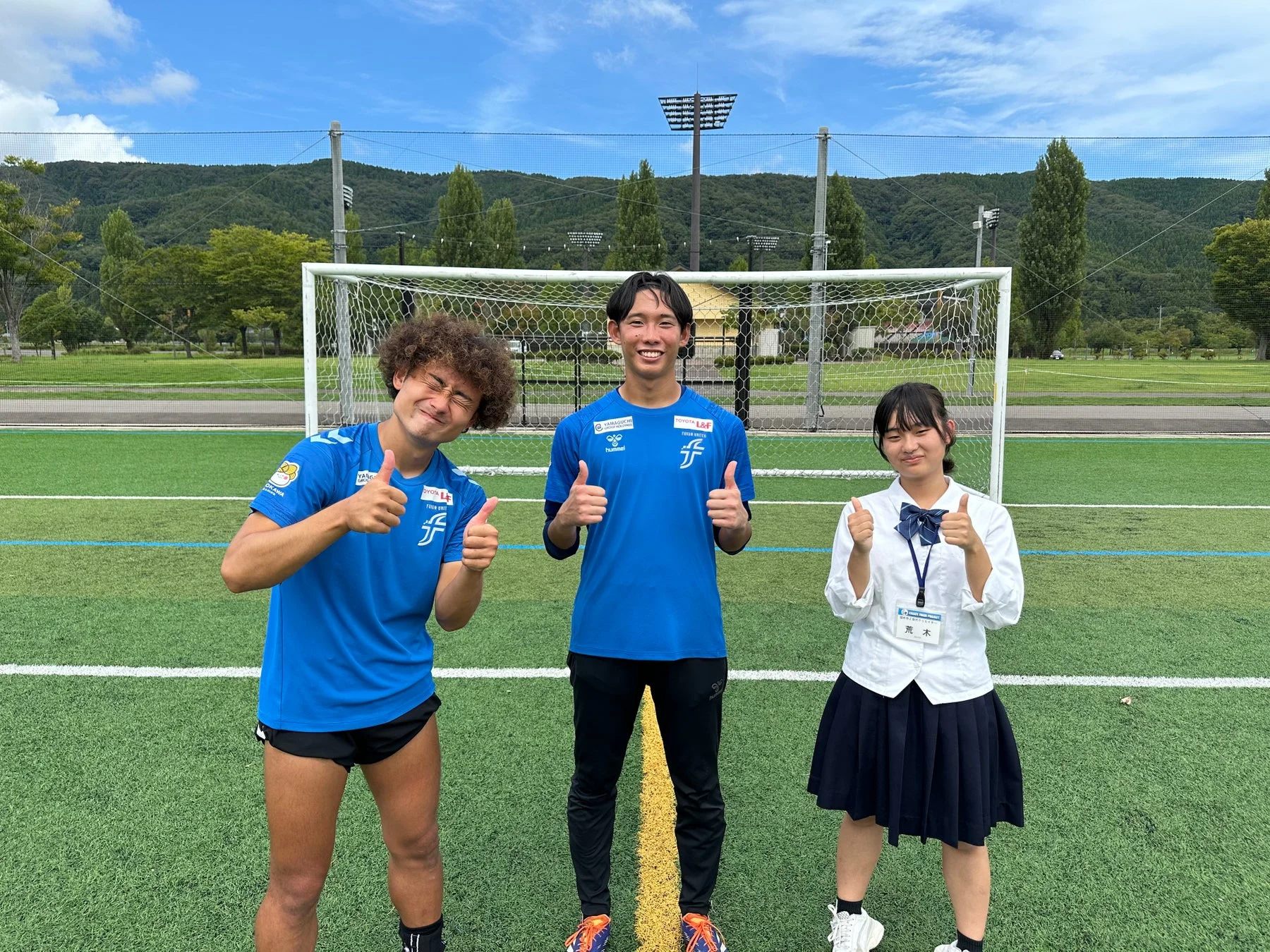 サッカー場で笑顔でサムズアップする3人の姿