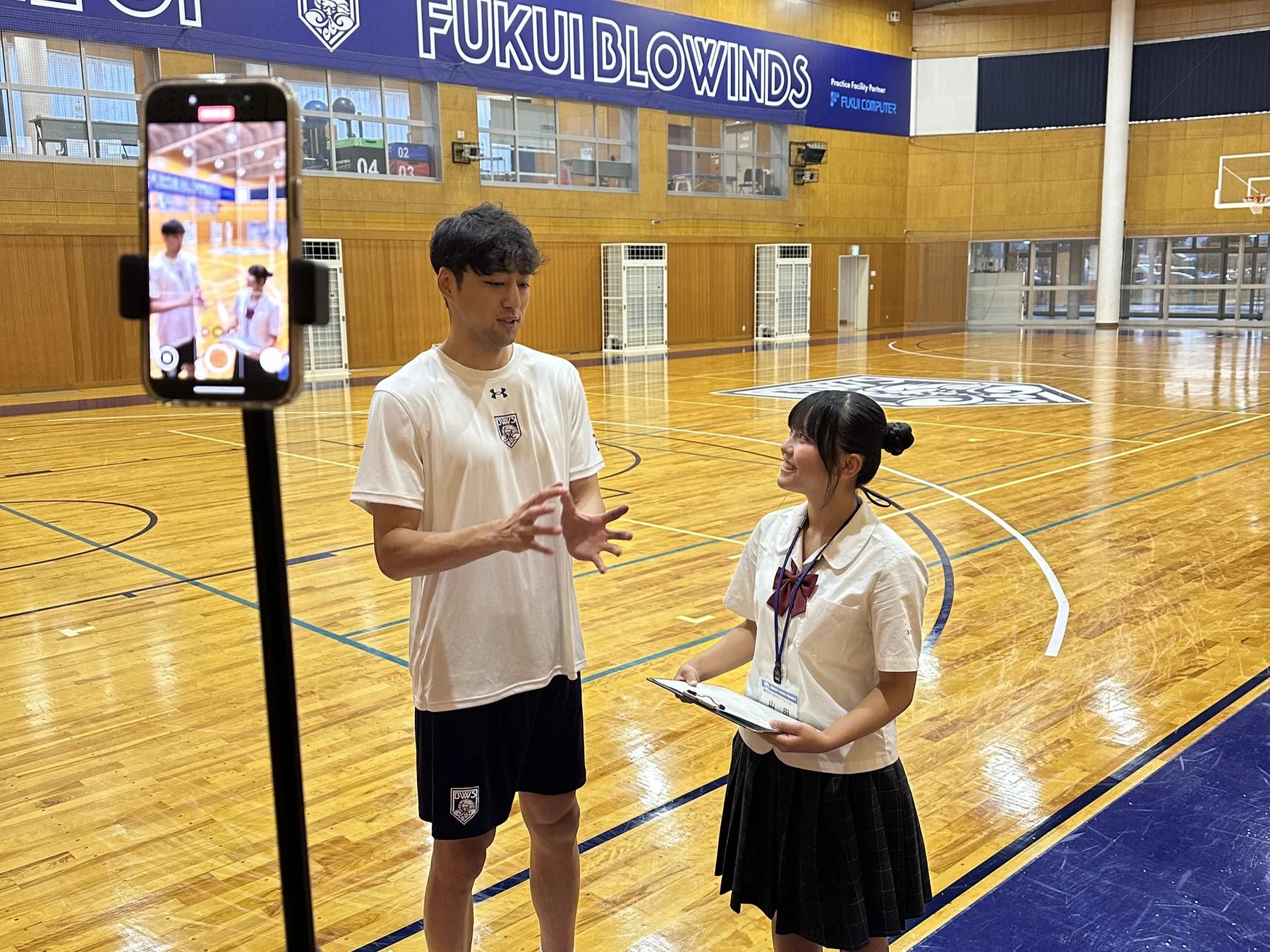 バスケットボールコートで男性アスリートが女子学生のインタビューを受けている様子