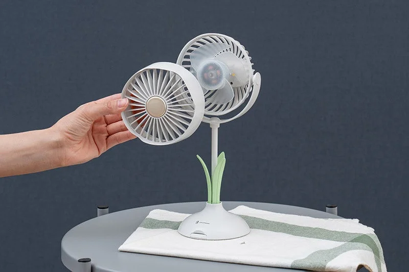 白い卓上扇風機を組み立てている様子です。片方のファン部分を手に持ち、植物のようなデザインのスタンドに取り付けようとしています。デスクやテーブルでの使用に適した、おしゃれな小型扇風機です。