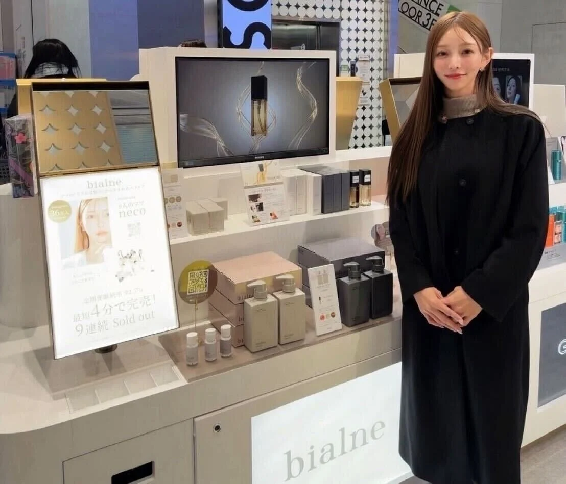 化粧品売り場で、黒いコートを着た女性が「bialne」製品のプロモーションディスプレイの横に立っています。