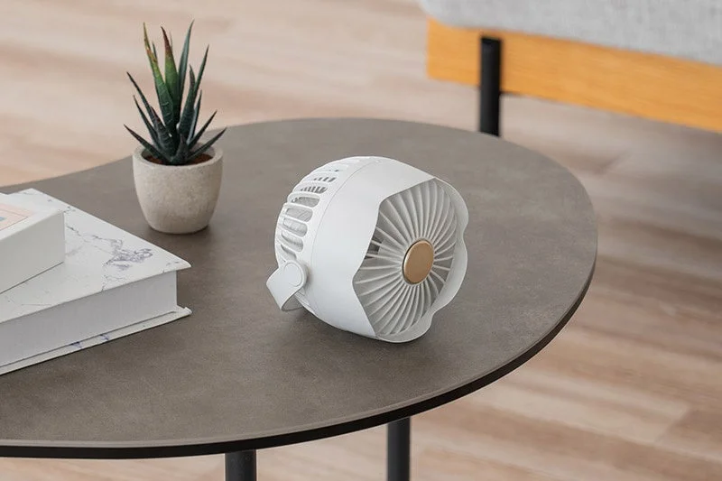 白い小型のポータブル扇風機が、観葉植物や本と一緒にコーヒーテーブルに置かれている。モダンで落ち着いた雰囲気の室内インテリアを示す画像です。