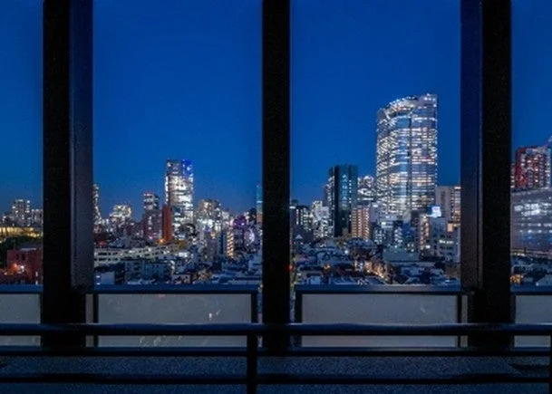 窓越しに広がる、高層ビル群が煌めく美しい都市の夜景です。深い青色の夜空の下、無数の光が輝き、都会的な雰囲気を醸し出しています。