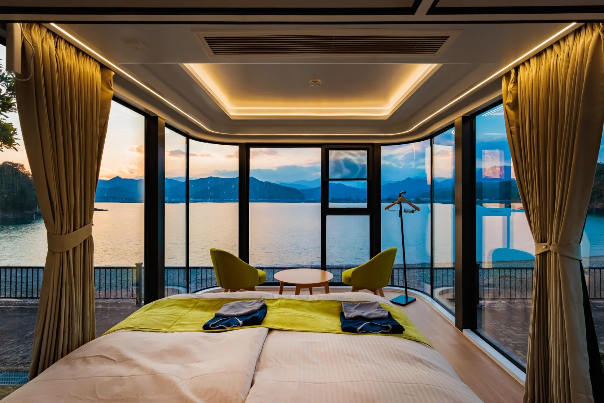 モダンで豪華なホテルの寝室から、湖または湾と山々が広がるパノラマの夕焼け景色