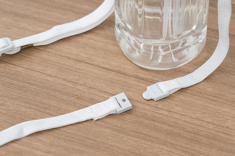木目調のテーブルに置かれた白いストラップが、プラスチック製のバックル部分で分離している様子を捉えた画像です。奥には透明なペットボトルの一部が見えます。ストラップの接続状態が強調されています。