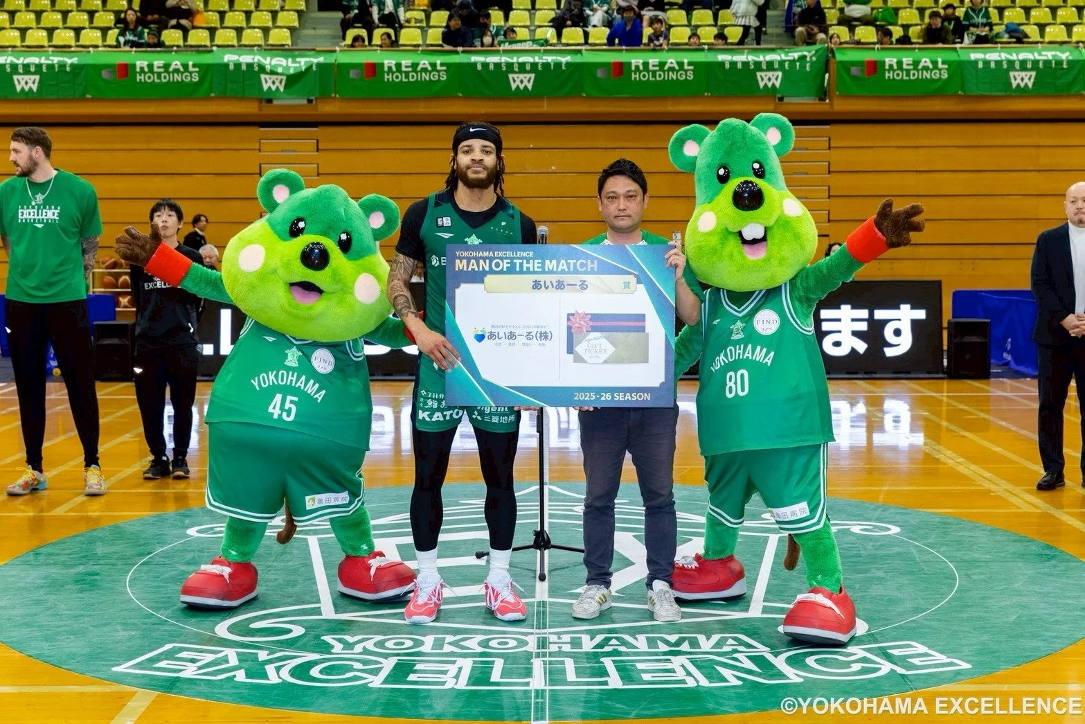 横浜エクセレンスのバスケットボール試合後、MVP選手と関係者、そして緑色のマスコット2体が「MAN OF THE MATCH」のボードを持って記念撮影をしている