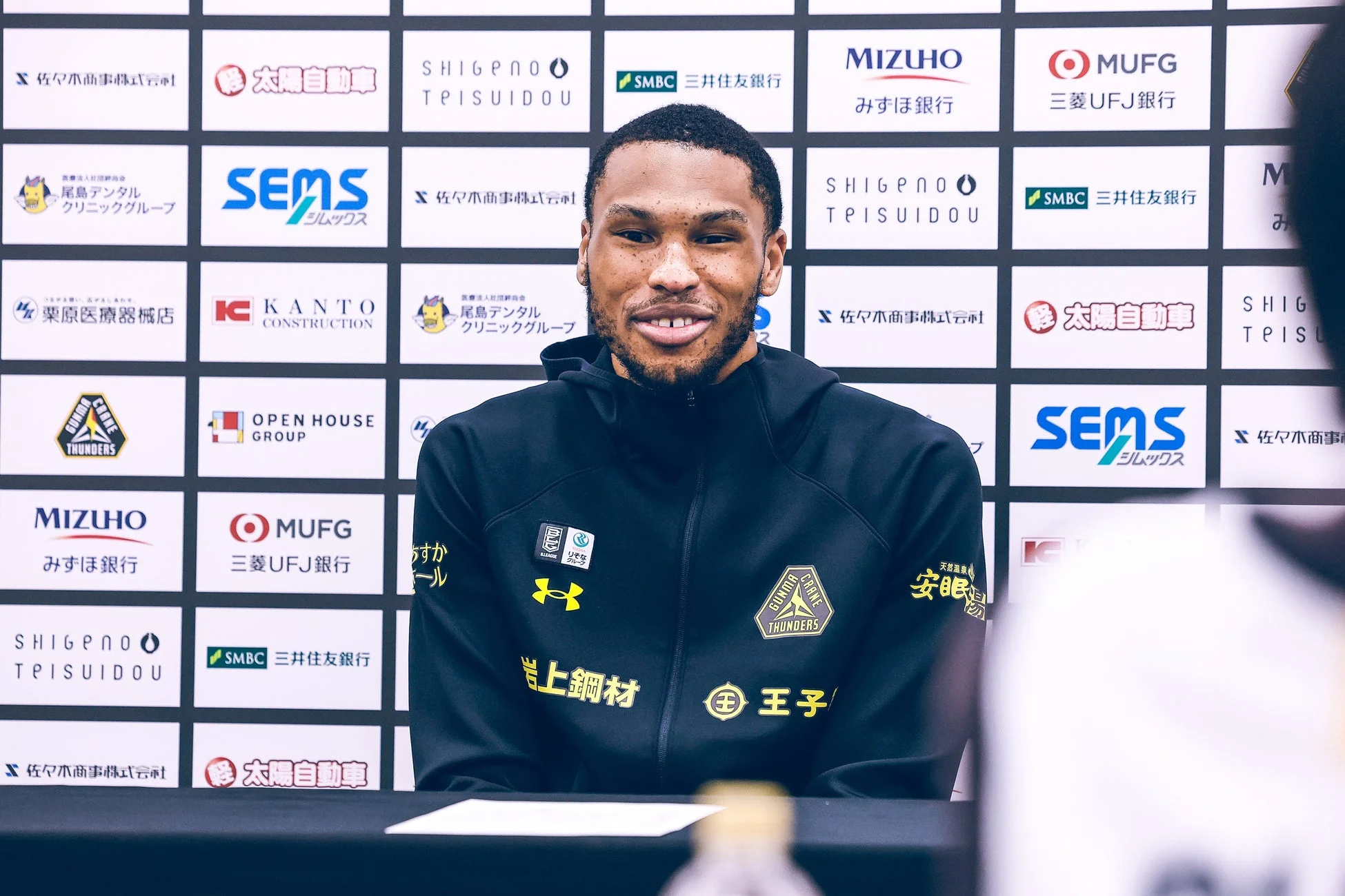 スポンサーロゴが並ぶ背景の前で座る群馬クレインサンダーズのケリー・ブラックシアー・ジュニア選手