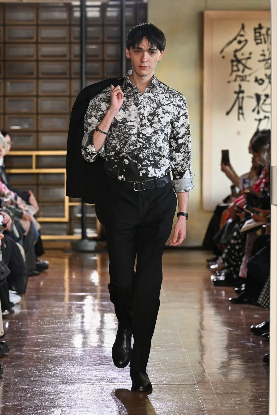 男性モデルがランウェイを歩いているファッションショーの様子です。白黒の柄シャツに黒いパンツを合わせ、ジャケットを肩にかけています。背景には書道アートが飾られています。