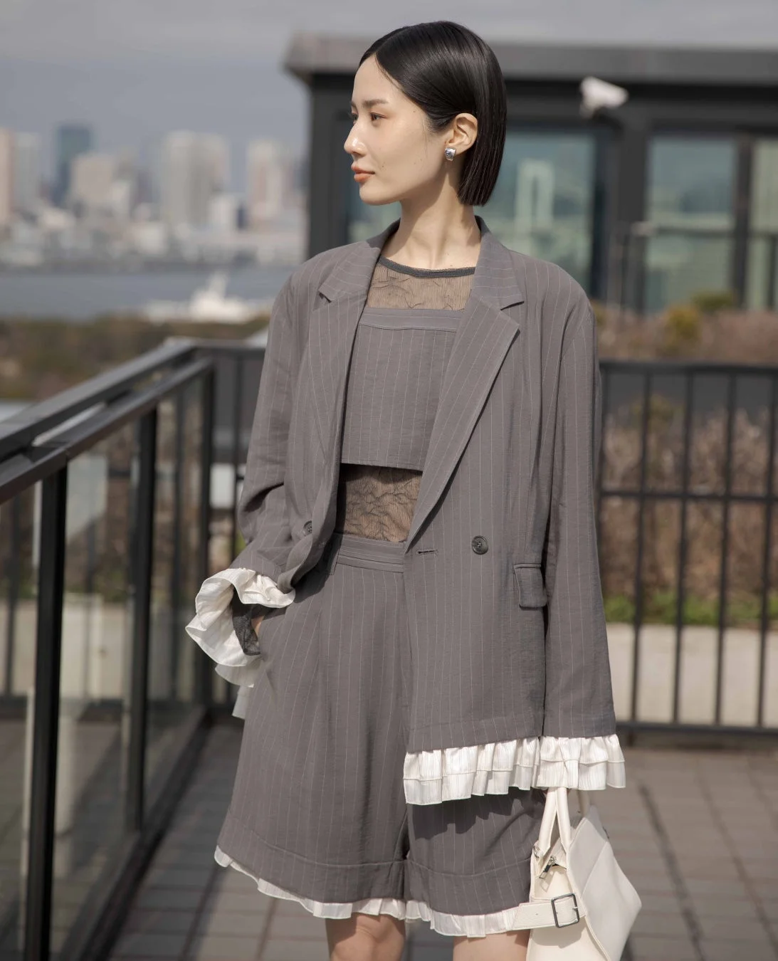 グレーのピンストライプのセットアップを着た女性が屋外でポーズをとっています。フリルが特徴的なデザインで、背景には都会の景色が広がっています。ショートヘアと上品なアクセサリーが洗練された印象を与えています。