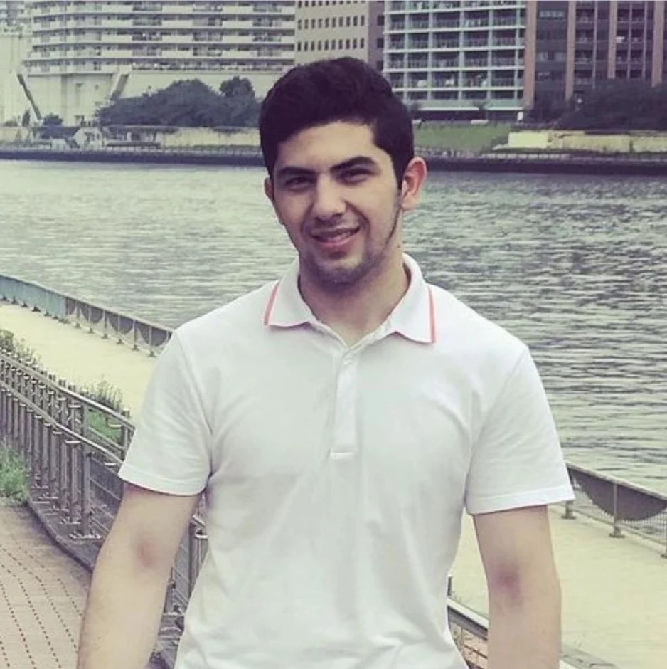 白いポロシャツを着た若い男性が、川沿いの歩道で笑顔を見せているポートレートです。背景には建物が立ち並ぶ都市の風景が広がっています。