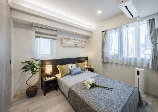 明るくモダンな寝室のインテリア写真です。ベッドには花が飾られ、両サイドにサイドテーブルとランプ、窓際には観葉植物が配置されています。快適で落ち着いた雰囲気の空間です。