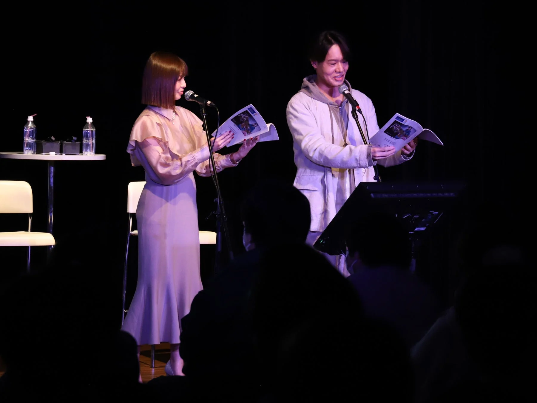 薄暗いステージ上でマイクを前に冊子を手に何かを話す男女