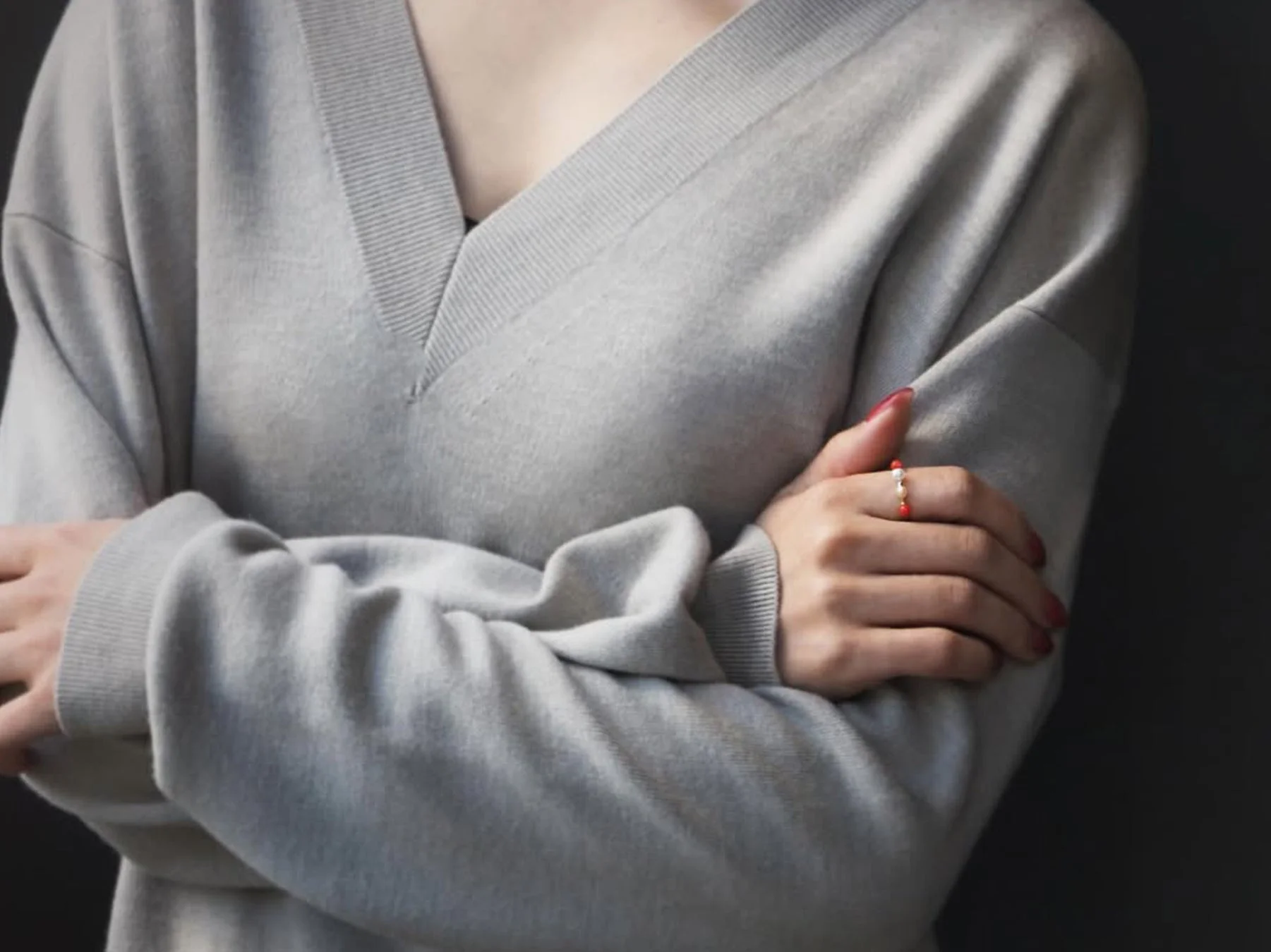 グレーのVネックセーターを着た女性が腕を組み、指には赤と白のビーズが付いたリングを身につけている