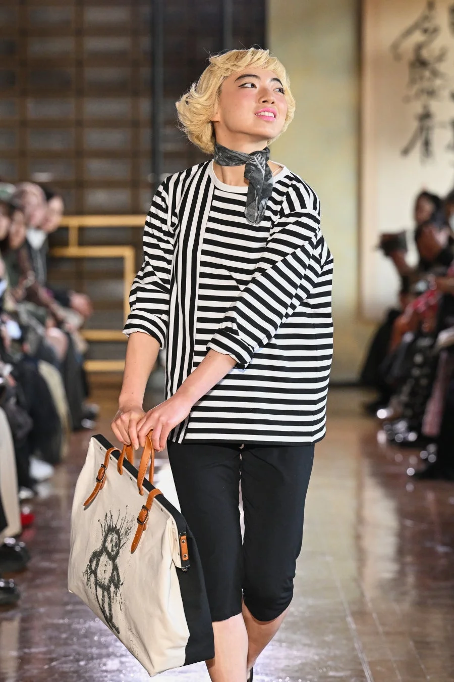 金髪の女性モデルが、黒と白のボーダー柄トップスと黒いカプリパンツを着用し、独特な絵柄のトートバッグを持ってランウェイを歩いています。背景には和風の建築と書道が見られ、ファッションショーの一場面を捉えた写真です。