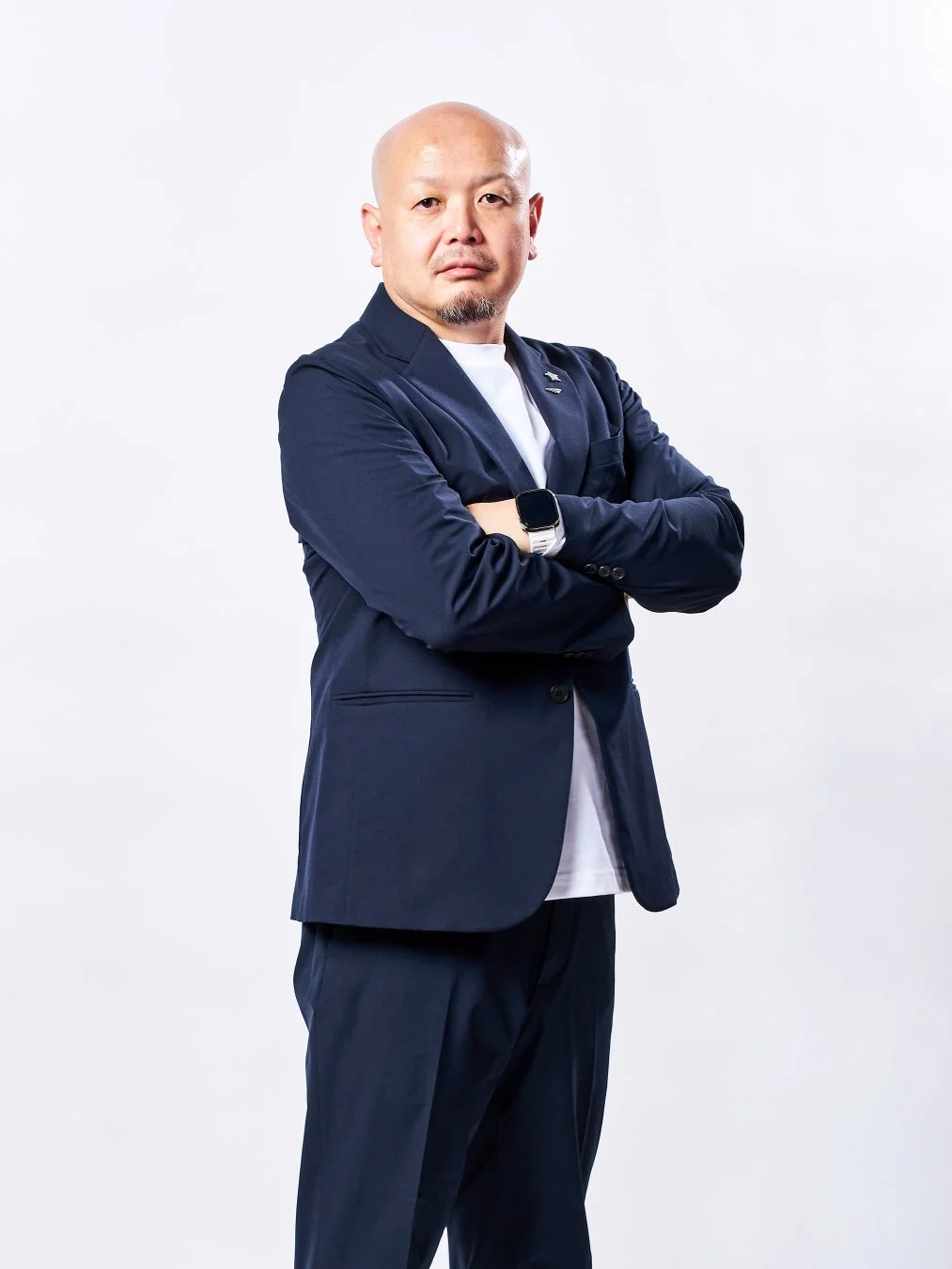 白い背景のスタジオで撮影された、紺色のスーツと白いTシャツを着た坊主頭の日本人男性のポートレートです。男性は腕を組み、カメラに向かって真剣な表情をしています。
