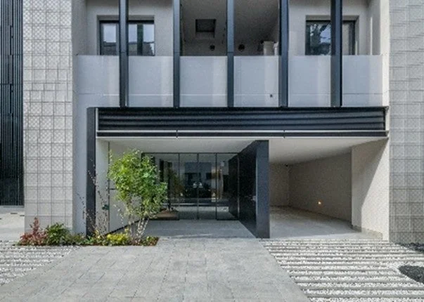 現代的でミニマリストなデザインの建物です。ガラス張りのエントランスと一体化したガレージが特徴で、すっきりとしたグレーのファサードとシンプルな植栽が都市的な雰囲気を醸し出しています。