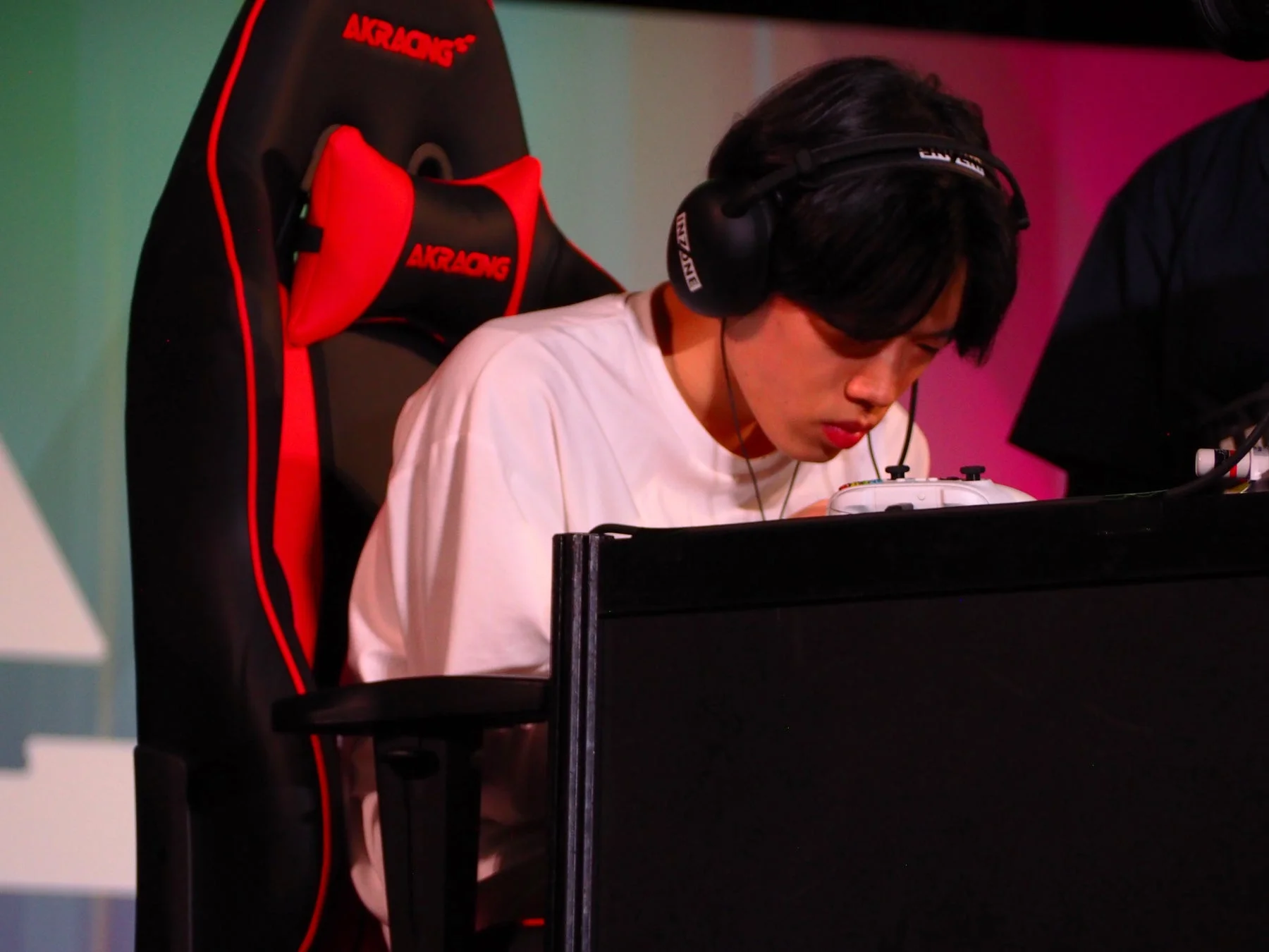 ゲーミングチェアに座り、ヘッドホンを装着した若い男性がゲームコントローラーを操作している様子。真剣な表情でゲームに集中しており、eスポーツイベントの一幕のようです。