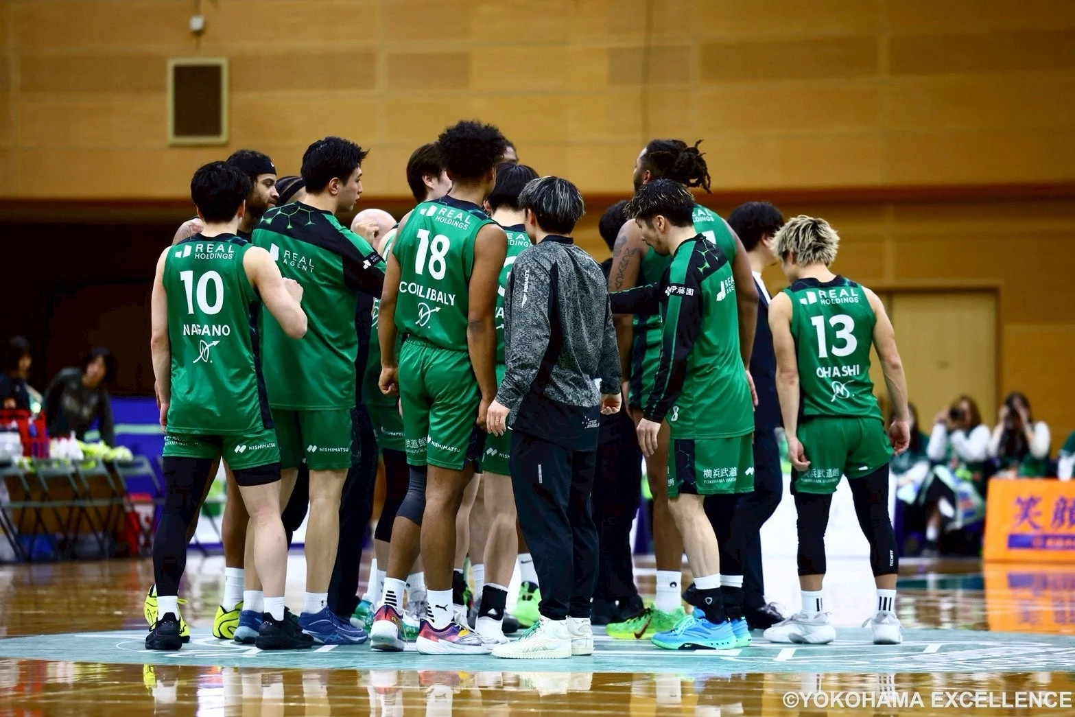 バスケットボールの試合中、緑色のユニフォームを着た選手たちがコート上で円陣を組んでいる