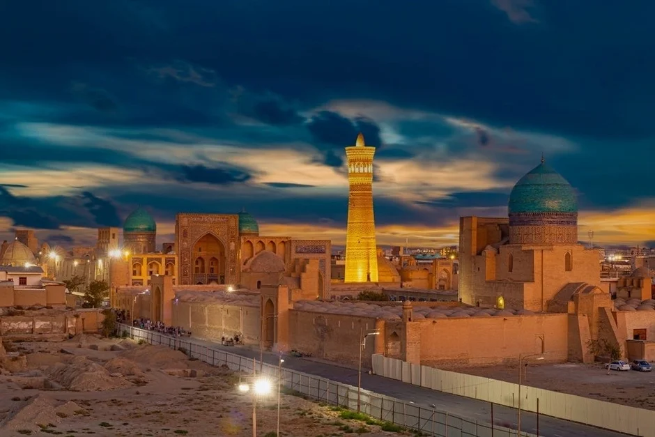 ウズベキスタンの古都ブハラにある歴史的な建造物が立ち並ぶ景観です。高くそびえるカリアンミナレットが特徴的で、夕暮れの空の下、建物が美しくライトアップされています。