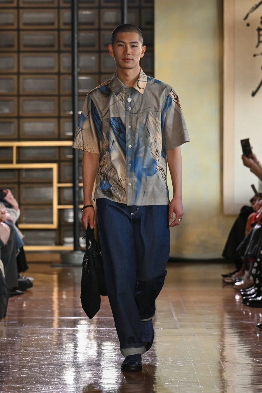 抽象的な青とベージュの柄の半袖シャツとワイドなデニムパンツを着用した若い男性モデルが、和風の背景のランウェイを歩いている様子。ファッションショーの一幕を捉えたスタイリッシュな画像です。