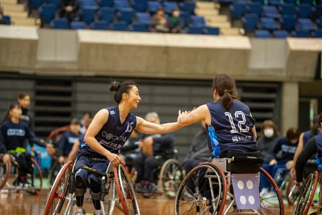 体育館で行われている車いすバスケットボールの試合中に、2人の女性選手が笑顔でハイタッチを交わしている瞬間