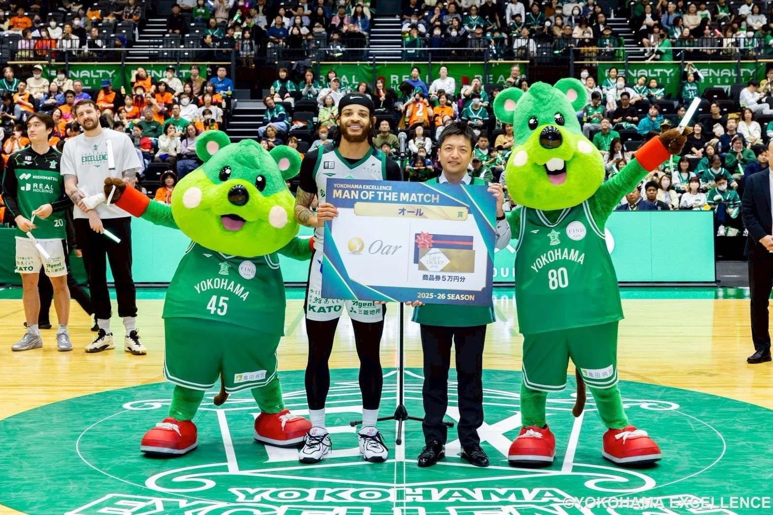 バスケットボールチーム「横浜エクセレンス」の試合後、選手がマン・オブ・ザ・マッチとして表彰されている。緑色の可愛らしいマスコットとスーツ姿の男性が、選手に「Oar」と書かれた賞品パネルを贈呈しており、アリーナには多くの観客がいる。