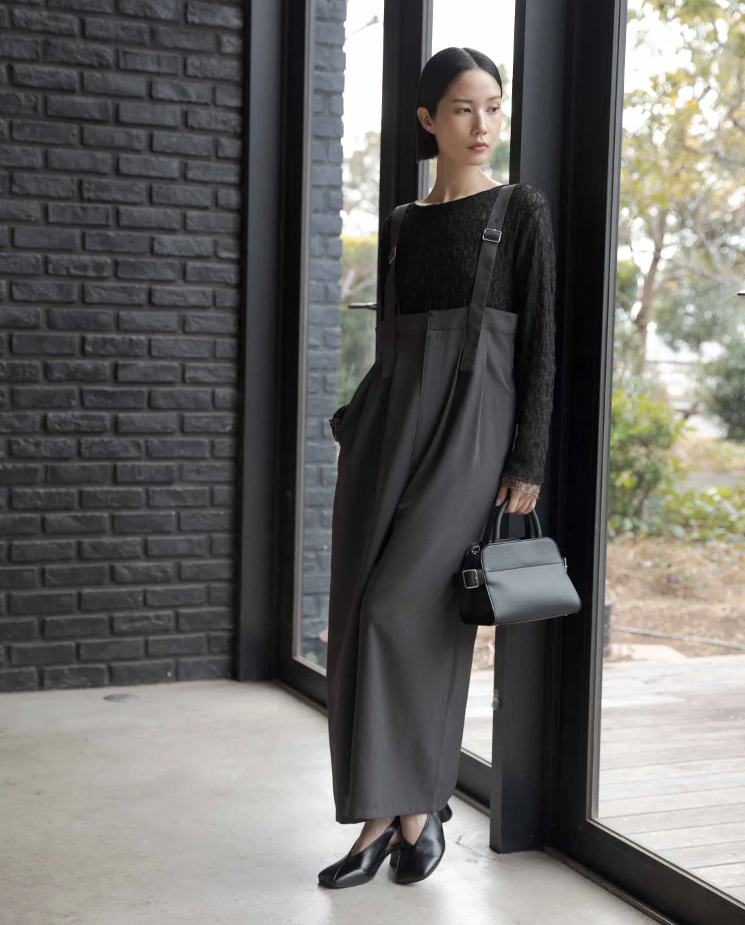 窓際に立つ女性の全身ショット。黒の長袖トップスにダークグレーのワイドサロペットを合わせた、シンプルながらもスタイリッシュなコーディネート。黒いハンドバッグとパンプスで全体を引き締め、モードな雰囲気を醸し出している。