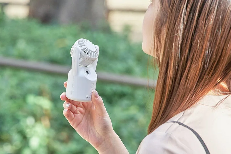 屋外で女性が白い携帯扇風機を使用している様子です。暑い日に涼を取るため、顔に向けて風を送っています。ミスト機能付きの可能性もあります。