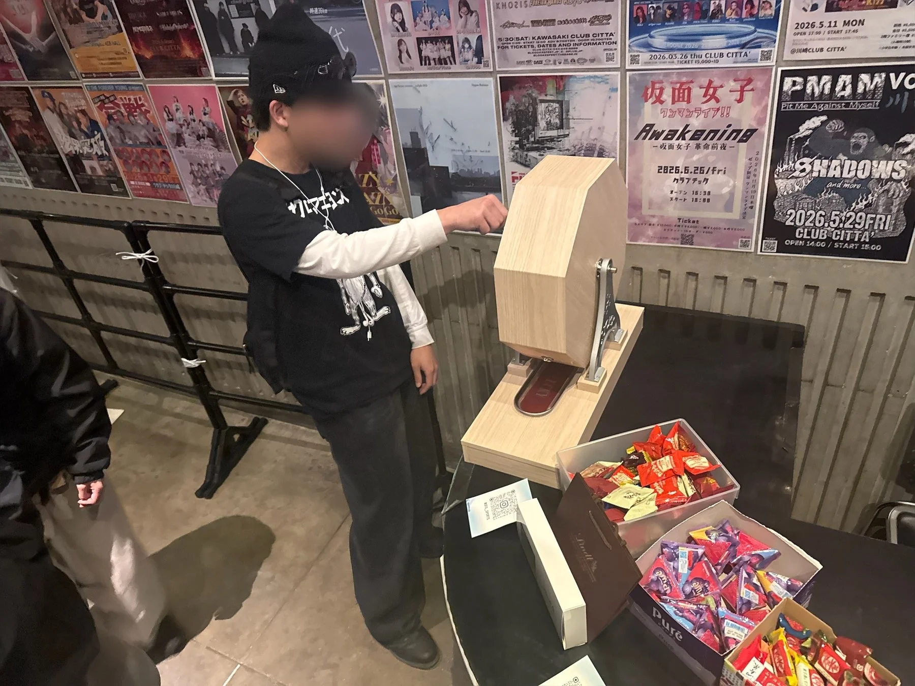 音楽イベント会場らしき場所で、一人の人物が木製の抽選機を回している。テーブルには景品と思われる様々なお菓子が置かれ、壁には「仮面女子」や「SHADOWS」などの公演ポスターが多数貼られている。