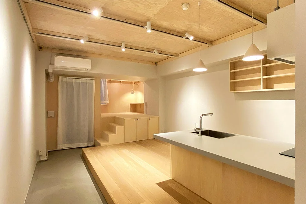 明るくモダンなアパートメントの室内。木目の天井と床が特徴的で、アイランドキッチンや段差のある収納スペースが配置されています。