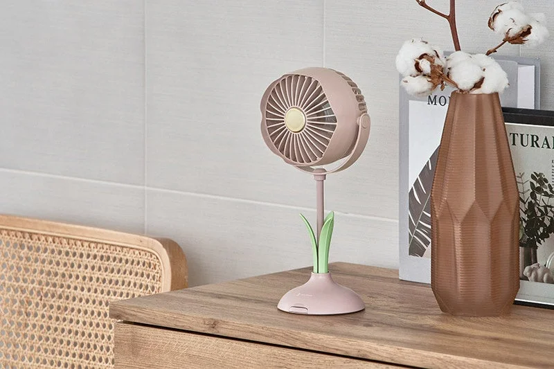 木製の家具の上に置かれたピンクの可愛らしい卓上扇風機と、ブラウンの花瓶に生けられたコットンフラワーが写っています。背景には白いタイル壁とラタンの椅子が見え、全体的にモダンでナチュラルな雰囲気のインテリアを捉えた画像です。