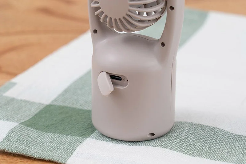 ベージュ色の携帯扇風機の底部をクローズアップした画像。USB充電ポートと小さなインジケーターライトが見え、緑と白のチェック柄の布の上に置かれている様子が捉えられています。