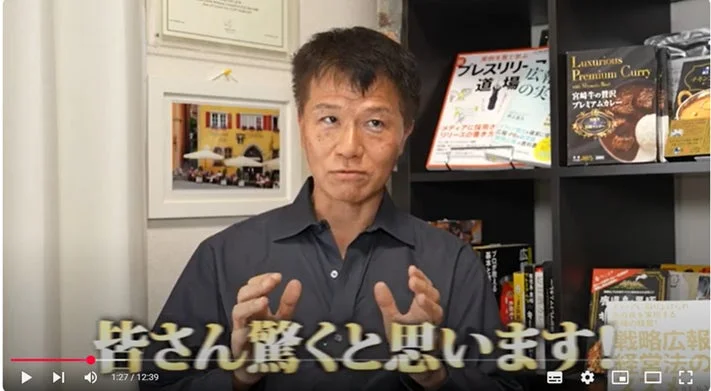 ビジネス系の動画の一場面で、男性が身振り手振りを交えて何かを説明している様子