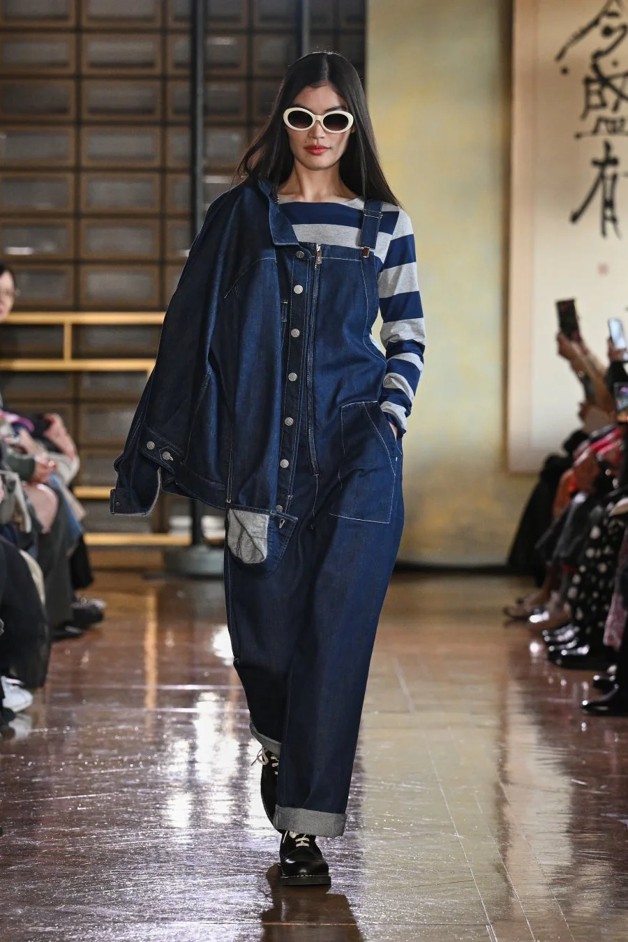 ランウェイを歩くモデルが写っています。ボーダーシャツの上にデニムのオーバーオールとジャケットを合わせたカジュアルな装いで、白いサングラスがアクセントになっています。背景には日本の伝統的な書道が見えます。