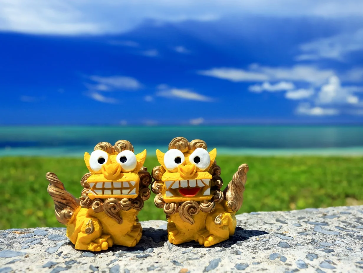 沖縄のエメラルドグリーンの海と青い空を背景に、愛らしい2匹の黄色いシーサーの置物が並んでいます。