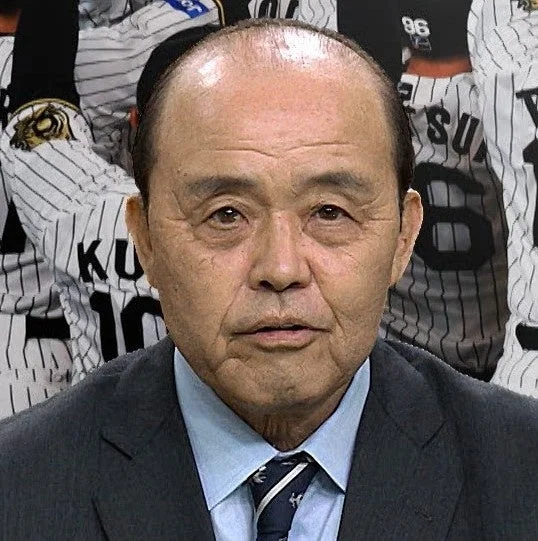 スーツ姿の日本人男性が真剣な表情で写っており、背景には野球のユニフォームを着た人々が見えます。野球関係者、おそらく監督かコーチのポートレートと考えられます。