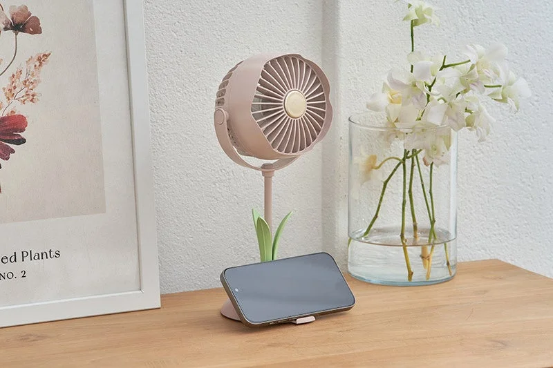 木製のデスク上に、チューリップ型の卓上扇風機、スマートフォン、白い花が生けられた花瓶、そして植物の絵が飾られたフレームが配置されており、おしゃれな室内空間を演出しています。