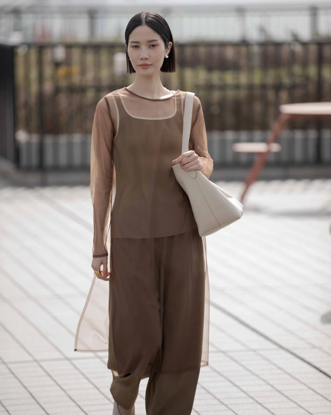 屋外のウッドデッキを歩くボブヘアの女性。ブラウンのシアー素材のトップスとワイドパンツを着用し、クリーム色のショルダーバッグを持っています。上品で洗練された印象です。