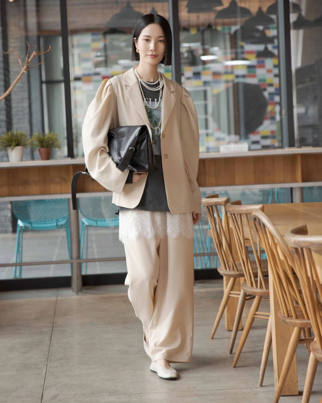 ベージュのジャケットとワイドパンツを着用したボブヘアの女性が、モダンなカフェのような空間でポーズをとっています。パールネックレスと黒いバッグがアクセントになっています。